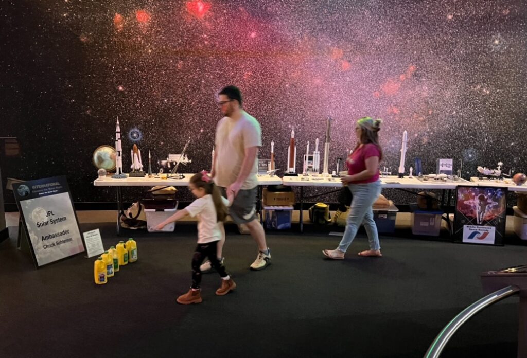 Family checking out the hands-on planetary gravity demonstration using milk jugs weighted as if on Earth, Moon, Mars, Phobos and Jupiter.